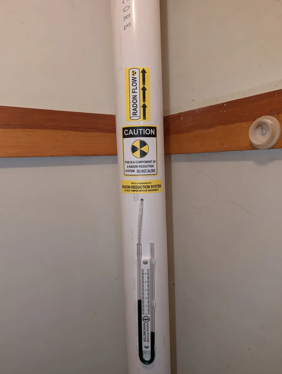 Radon vent pipe with warning labels and a U-tube manometer showing the system pulling suction