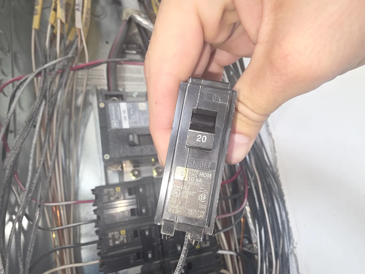 A pulled 20 amp breaker held up in front of an open panel full of older wiring